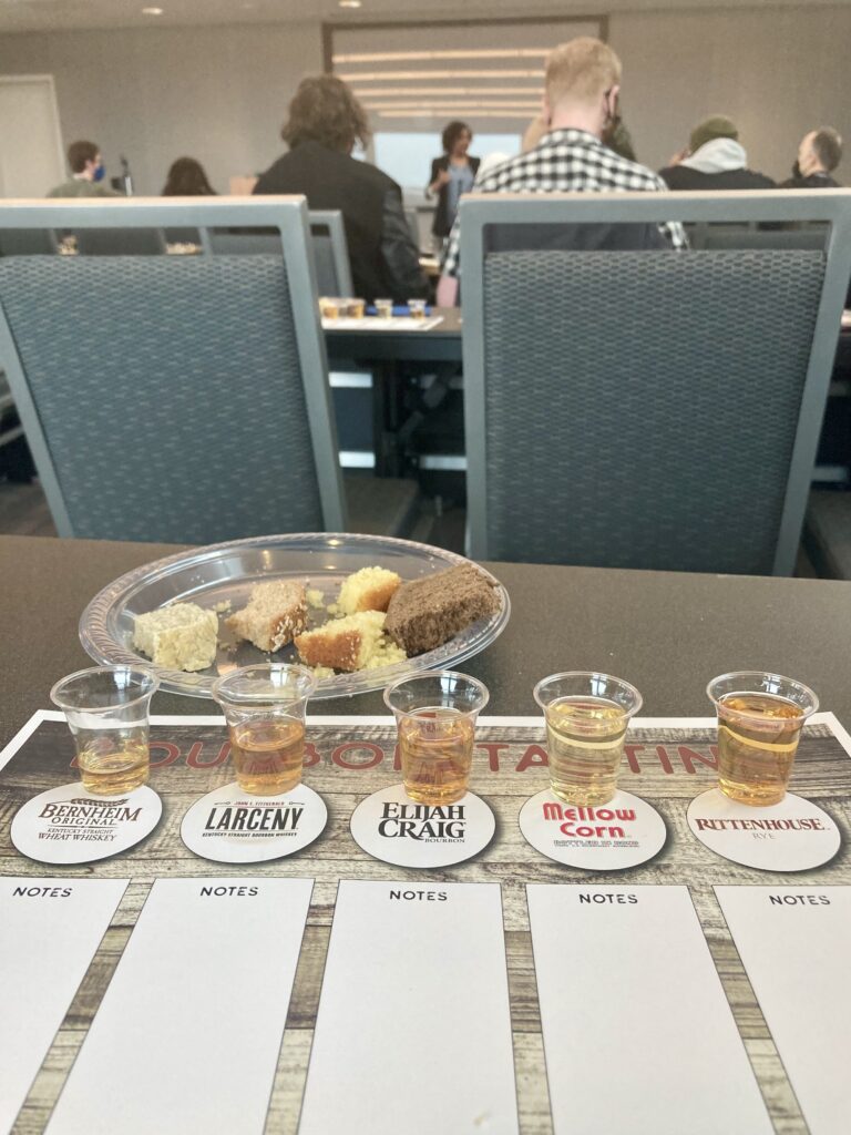 A plate with different types of bread and a tasting mat with labels under five small plastic cups of whiskey from Heaven Hills Distillery. 