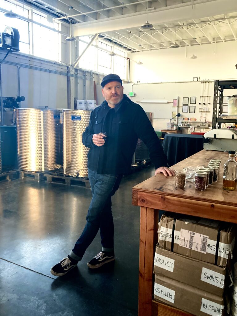 Adam Fream, the Sales Manager at Fast Penny Spirits, talking and leaning against a table with herbs and botanicals during our tour of Fast Penny Spirits. 