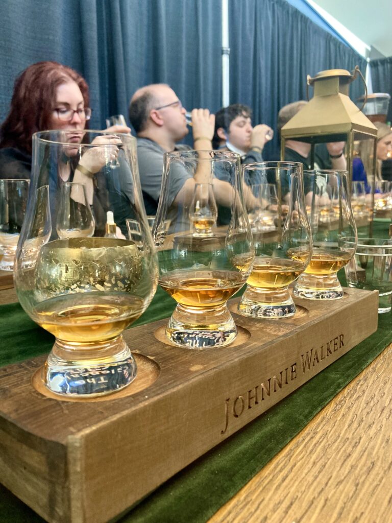 Four, tasting glasses with whiskey in front of people drinking whiskey in the background in the Johnnie Walker booth at the Carnival of Cocktails during Seattle Cocktail Week.