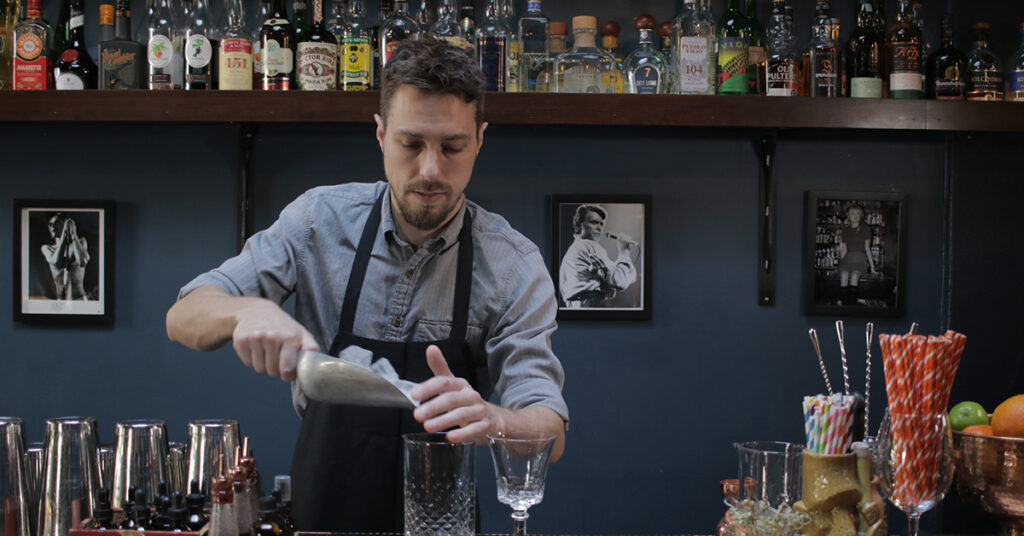 Tom Lindstedt, General Manager at La Moule in Portland, Oregon, preparing a cocktail behind the bar, pouring ice into a mixing glass.